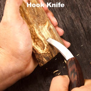 以下が含まれることがあります： 曲がった刃のフックナイフを使って木材を彫刻しています。ナイフは濃い木製の柄と銀色の刃を持っています。「Hook Knife」という文字が画像上部に白文字で表示されています。