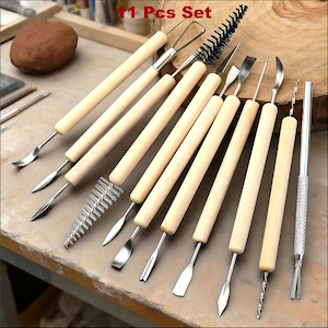 以下が含まれることがあります： 木製のハンドルとステンレス鋼の先端が付いた、11ピースの陶器ツールセット。このセットには、粘土を彫刻、成形、詳細化するためのさまざまなツールが含まれています。ツールは木製の表面に配置されています。
