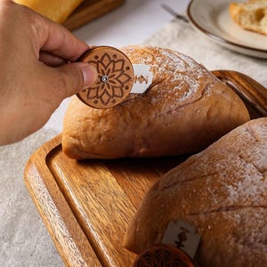 Könnte beinhalten: Ein Brotmesser aus Holz mit einem dekorativen Blumendesign wird in der Nähe eines Brotlaibs gehalten. Das Brot liegt auf einem Holzschneidebrett. Eine Rasierklinge ist in das Brot eingesetzt. Das Brot ist hellbraun und mit Mehl bestäubt.