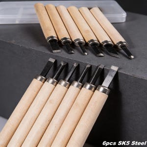 May include: A set of wood carving tools with light wooden handles and black steel blades. The tools are arranged in two rows, with a clear plastic case in the background. The text "6pcs SK5 Steel" is visible at the bottom.