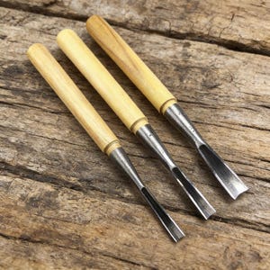 Peut inclure: Trois ciseaux à bois avec des manches en bois de couleur claire et des lames en métal argenté. Les ciseaux sont disposés en diagonale sur une surface en bois vieilli, mettant en valeur leurs bords tranchants et leur savoir-faire. Les manches sont cylindriques.