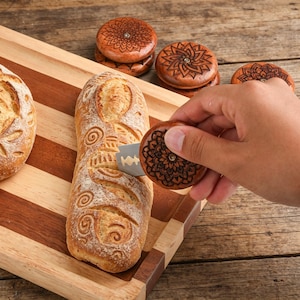 May include: A wooden bread lame with a decorative mandala design is held near a loaf of bread on a wooden cutting board. The bread has intricate scoring patterns. Several other wooden lames with mandala designs are also visible.
