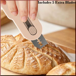 May include: A beige bread lame with a silver blade is used to score a loaf of bread. The bread has a decorative pattern cut into the top. The text "Includes 5 Extra Blades" is visible in the image.