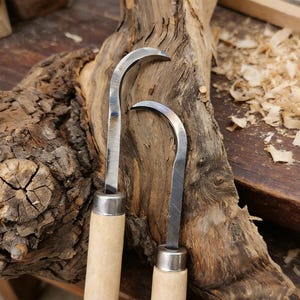 Puede incluir: Dos cuchillos de tallado de metal con hojas curvas y mangos de madera clara. Las hojas son plateadas y los mangos de color beige claro. Los cuchillos se colocan sobre un fondo de madera rugosa y texturizada.