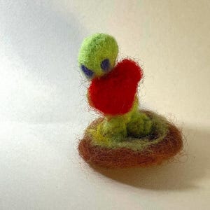 May include: A handmade felt alien figure in shades of green, purple, and red. The alien is holding a red heart and is nestled in a brown nest. The figure is a whimsical, handcrafted item.