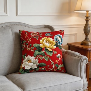 May include: A square decorative pillow with a vibrant red background. The pillow features a floral pattern with large, detailed flowers in white, yellow, and blue, along with green leaves and stems. The pillow is placed on a gray sofa.