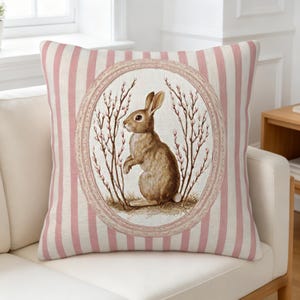 May include: Square decorative pillow with a pink and white striped background. The center features a framed illustration of a brown rabbit standing among branches. The pillow is on a white couch.