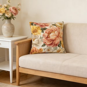 May include: A decorative square pillow with a large floral pattern featuring peach, yellow, and white flowers. The pillow is placed on a beige sofa with wooden arms. A vase of flowers sits on a white side table.