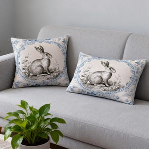 Puede incluir: Dos cojines decorativos con una ilustración vintage de conejo. Los cojines presentan un fondo crema con un borde floral azul. El conejo está representado en un dibujo detallado en escala de grises, añadiendo un toque de encanto clásico a la decoración del hogar.