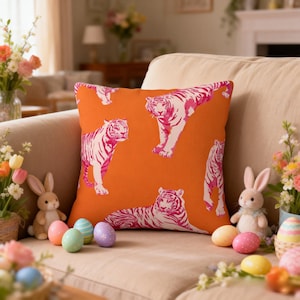 May include: Square orange cushion featuring a repeating design of pink and white tigers. The cushion is on a beige sofa, with Easter decorations such as painted eggs and toy rabbits nearby.
