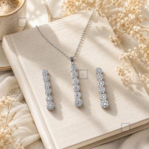 Peut inclure: Une parure de bijoux argentée composée d'un collier et d'une paire de boucles d'oreilles assorties. Chaque pièce présente un agencement vertical de cinq grappes circulaires de petites pierres claires et scintillantes sur monture.