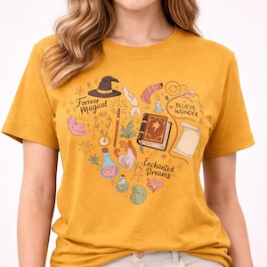 Op de afbeelding: Mosterdgeel T-shirt met een hartvormig ontwerp met magische elementen. Het ontwerp bevat een heksenhoed, een toverboek, een toverstaf en de zinnen "Forever Magical", "Believe in Wonder" en "Enchanted Dreams".