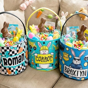 Puede incluir: Tres cestas de Pascua llenas de golosinas están sobre un sofá beige. Cada cesta tiene un diseño y un nombre diferentes: Roman, Connor y Braxtyn. Las cestas contienen conejos de chocolate, caramelos y hierba decorativa. Una guirnalda de zanahorias y conejos cuelga encima.