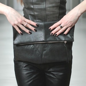 May include: A strapless black leather top with vertical seam detailing and a matching black leather fold-over clutch bag featuring a horizontal metal zipper closure. The ensemble is paired with black leather trousers and dark sunglasses.