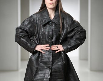 May include: A black leather coat featuring a patchwork design with visible seams, a wide collar, and a front button closure. The coat has long sleeves with buttoned cuffs and a knee-length hemline, constructed from multiple panels of black leather.