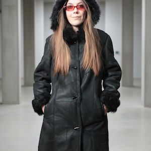 May include: A black long-length coat featuring a hood with black faux fur trim, long sleeves with black faux fur cuffs, and a front button closure with toggle-style fasteners. The coat is constructed from multiple panels of black material.