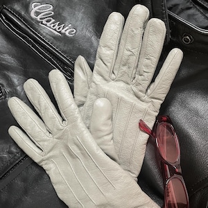 May include: A pair of light gray leather gloves with three vertical stitched lines on the back of the hand, resting on a black leather jacket with the word Classic embroidered in white thread, accompanied by a pair of dark red tinted sunglasses.