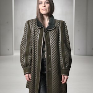 May include: An olive green coat featuring a prominent chevron-patterned texture across the front and sleeves. The garment includes a dark faux fur collar, long sleeves with cuffs, and a front button closure. The design incorporates vertical panels.