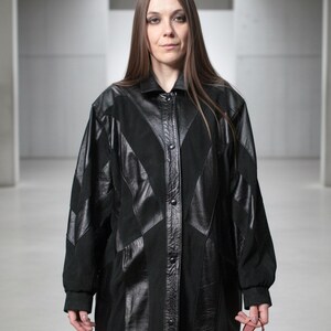 Peut inclure: Long manteau noir avec un devant boutonné et un design géométrique unique. Le manteau présente un mélange de textures, avec des panneaux en cuir et en daim créant un contraste visuel saisissant. La personne tient des lunettes de soleil rouges.