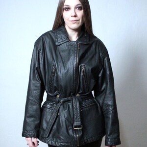 May include: A black leather jacket featuring a fold-down collar, a full-length front zipper, and two zippered chest pockets. The jacket includes a matching leather belt with a metal buckle at the waist and long sleeves with stitched detailing.