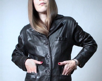 Puede incluir: Una chaqueta de cuero negro con cuello, mangas largas y cierre frontal de botones. La prenda incluye dos grandes bolsillos de parche en la cintura y un pequeño bolsillo en el pecho, todo confeccionado en material de cuero negro liso.