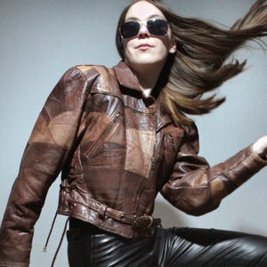 Könnte beinhalten: Eine braune Patchwork-Lederjacke mit Kragen, diagonalen Reißverschlusstaschen und seitlichen Schnürdetails. Die Jacke verfügt über einen Taillengürtel mit Schnalle und wird mit einer schwarzen Lederhose kombiniert. Das Kleidungsstück hat ein braunes Finish.