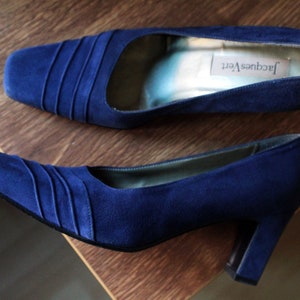 May include: A pair of deep blue suede pumps featuring a square toe with subtle pleated detailing across the vamp. The interior of the shoe is lined in a light-colored material with a visible Jacques Vert brand label on the heel insole.