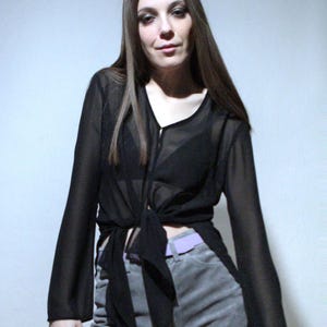 May include: A black sheer long-sleeve blouse with a front tie closure and a V-neckline, paired with grey shorts featuring a light purple belt. The blouse has wide, flowing sleeves and is worn over a black undergarment, creating a layered appearance.