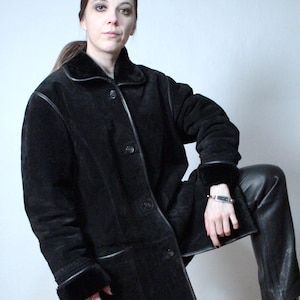 May include: A black suede coat featuring a fold-over collar, long sleeves with cuffs, and a button-front closure. The coat has smooth leather trim along the seams, collar, and cuffs, with a soft interior lining visible at the edges of the garment.