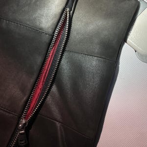May include: A dark charcoal gray leather panel featuring a vertical silver metal zipper with a vibrant red fabric lining. The leather surface displays a smooth texture with visible stitched seams, positioned next to a sewing machine presser foot.