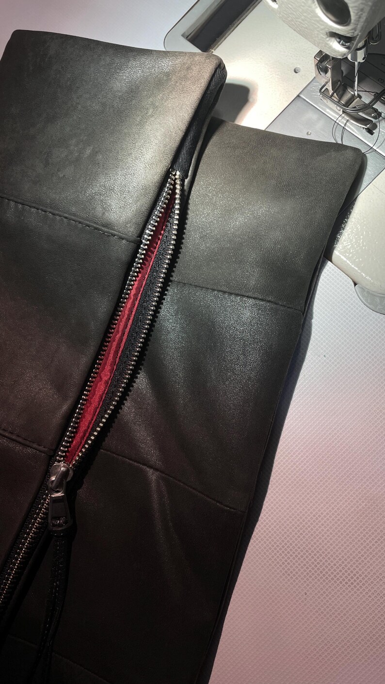 May include: A dark charcoal gray leather panel featuring a vertical silver metal zipper with a vibrant red fabric lining. The leather surface displays a smooth texture with visible stitched seams, positioned next to a sewing machine presser foot.