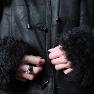 May include: A black leather coat with a hood and fur-lined cuffs. The coat has a button closure and is being fastened. The coat appears to be warm and suitable for cold weather.