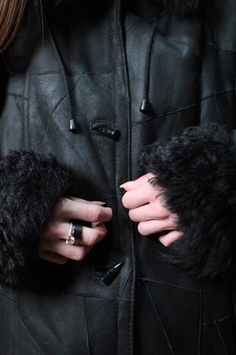 May include: A black leather coat with a hood and fur-lined cuffs. The coat has a button closure and is being fastened. The coat appears to be warm and suitable for cold weather.