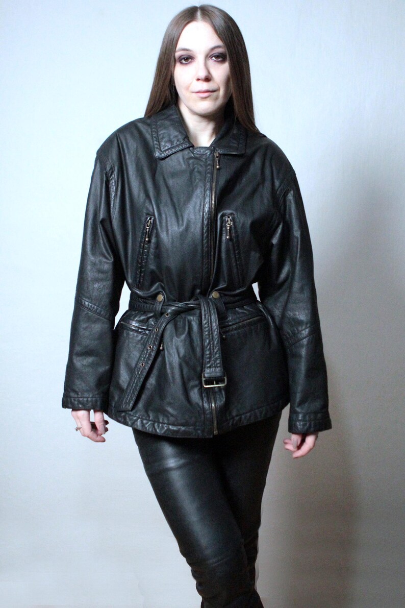 May include: A black leather jacket featuring a fold-down collar, a full-length front zipper, and two zippered chest pockets. The jacket includes a matching leather belt with a metal buckle at the waist and long sleeves with stitched detailing.