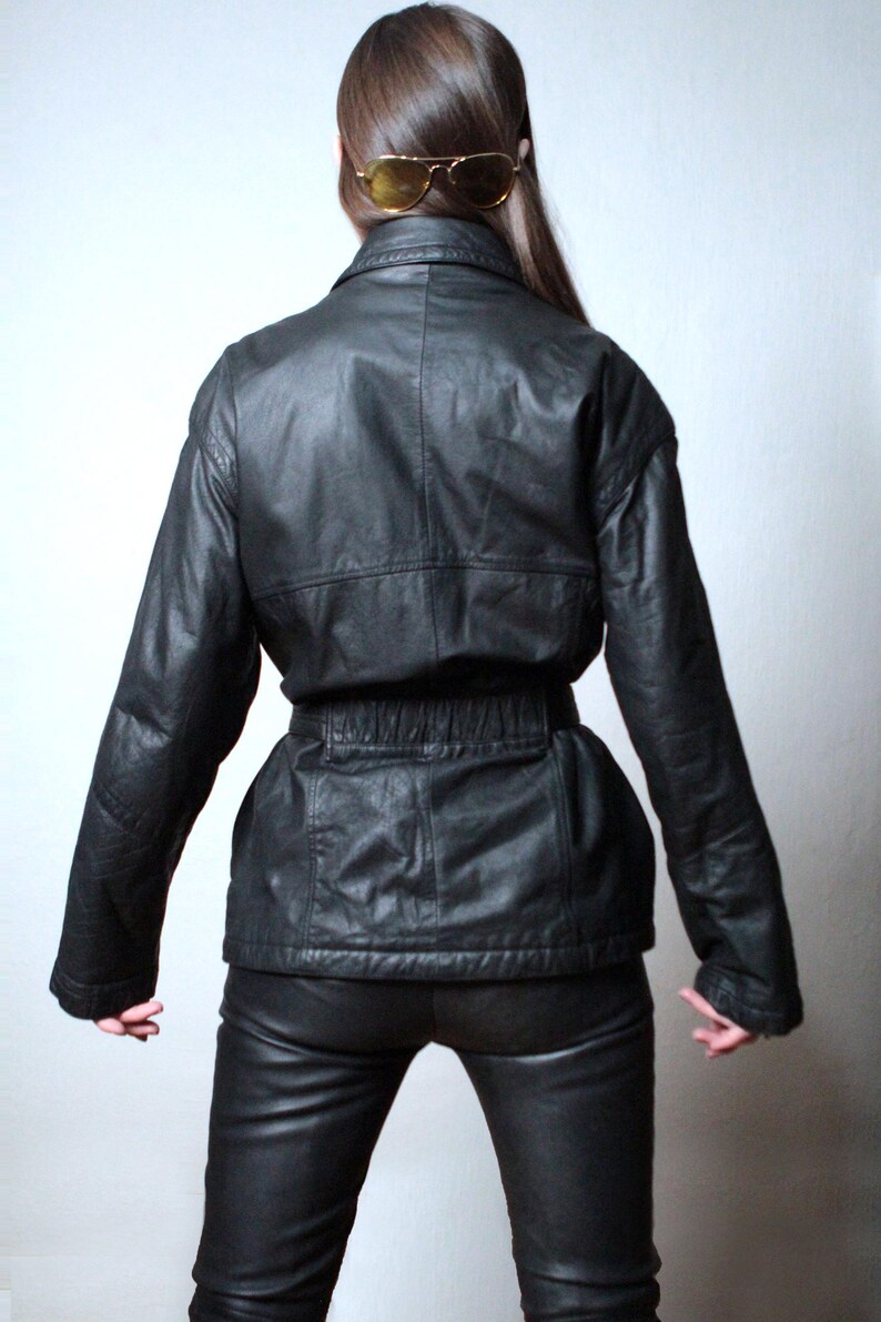 May include: A black leather jacket featuring a collared neckline, long sleeves, and a cinched waist with an elasticized band. The jacket has a smooth texture and visible seam detailing across the back and shoulders, paired with matching black leather pants.
