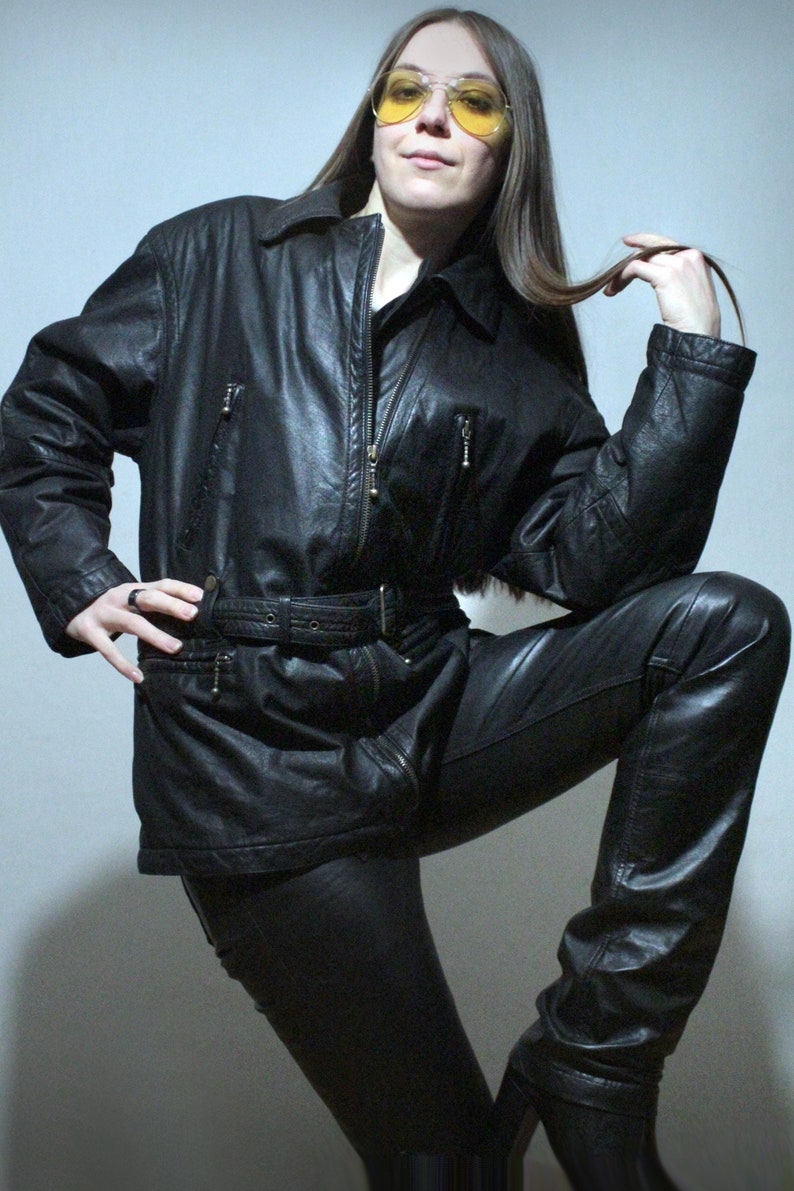 May include: A black leather jacket with a front zipper, a matching belt at the waist, and two zippered chest pockets. The jacket features a classic collar and long sleeves, paired with matching black leather trousers and yellow-tinted aviator glasses.