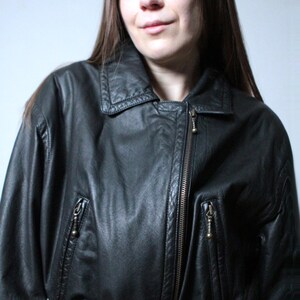 May include: A black leather jacket featuring a pointed collar, an asymmetrical front zipper closure, and two zippered pockets on the chest. The jacket includes a matching leather belt at the waist with metallic buttons and long sleeves.