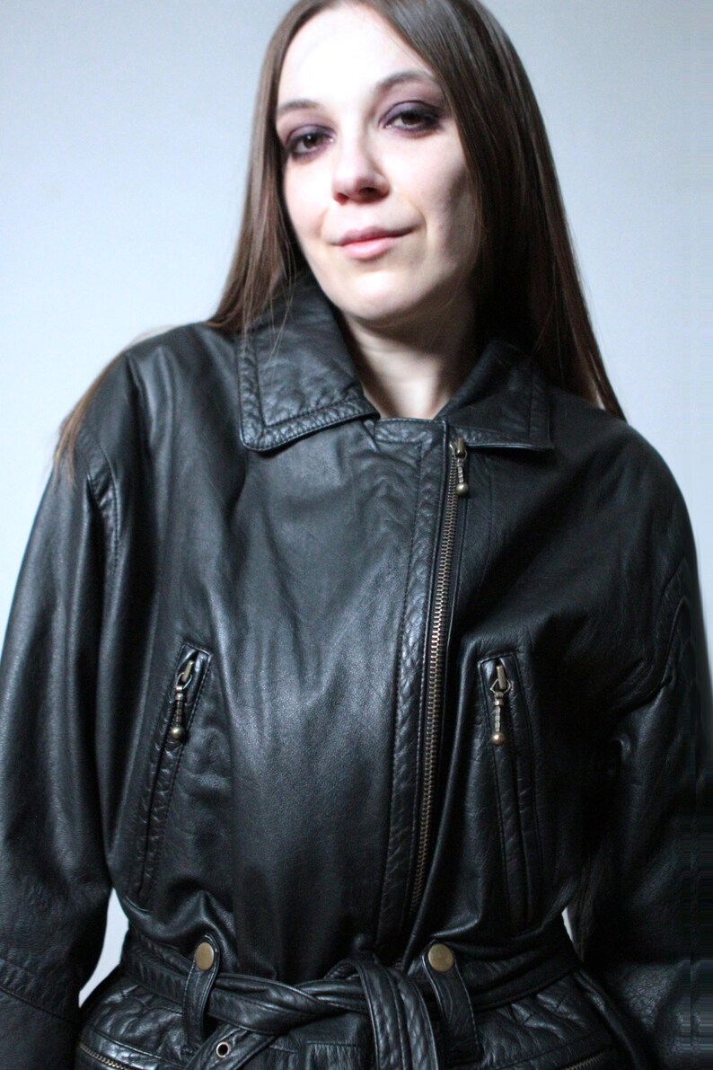 May include: A black leather jacket featuring a pointed collar, an asymmetrical front zipper closure, and two zippered pockets on the chest. The jacket includes a matching leather belt at the waist with metallic buttons and long sleeves.