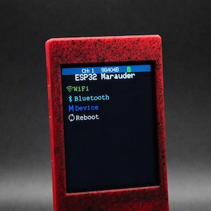 Peut inclure: Un appareil électronique rouge avec une finition texturée et un écran rectangulaire affichant le texte ESP32 Marauder. L'interface présente un menu avec les options WiFi, Bluetooth, Device et Reboot, ainsi qu'un indicateur de canal et une icône.