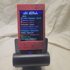 May include: A black and red WiFi sniffer device with a screen displaying network analysis options. The device is mounted on a black stand. The screen shows text including "WiFi Sniffers" and several scan options.