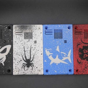 May include: Four rectangular cases with distinctive designs. The cases are black, white, blue, and red, each with a splatter paint effect. Designs include a butterfly, spider, shark, and a tribal-style face.