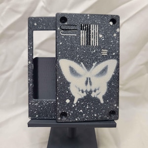 May include: A black and white decorative object with a butterfly skull design. The object has a speckled pattern and is mounted on a black stand. The butterfly skull design is white.