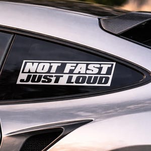 Peut inclure: Une voiture de sport argentée avec un autocollant noir sur la vitre qui dit "NOT FAST JUST LOUD" en lettres blanches en gras. La voiture a un aileron noir et un intérieur rouge visible à travers la vitre.