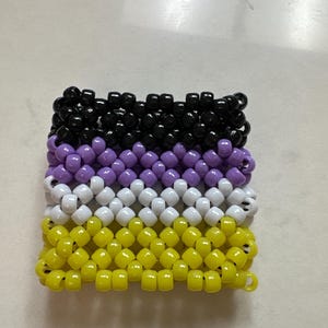 May include: A square-shaped beaded bracelet featuring the non-binary pride flag colors: black, purple, white, and yellow. The beads are arranged in horizontal rows, creating a textured surface. The bracelet is likely handmade.