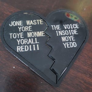 Puede incluir: Un objeto negro en forma de corazón, dividido en dos, con texto blanco a cada lado. El lado izquierdo dice "JONE WASTE YORE TOYE MONME YORALL REDIII" y el lado derecho dice "THE VOICE INSOIDE MOYE YEDD".