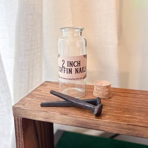May include: A clear glass jar with a label reading "2 INCH COFFIN NAILS" sits on a wooden surface. Two dark metal coffin nails and a cork stopper are next to the jar. The website theimmortalafterlife.com is printed on the label.