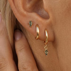 May include: A set of gold-toned jewelry consisting of two small hoop earrings and one stud earring. The stud and one hoop feature a small, marquise-cut, olive-green gemstone. The jewelry is arranged in a vertical line along the earlobe.