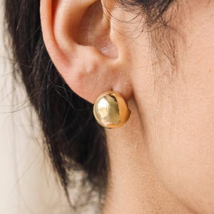 May include: A polished gold-tone stud earring featuring a smooth, rounded, dome-shaped design. The earring has a reflective, metallic surface and is secured to the earlobe with a post-back mechanism, presenting a simple and clean circular silhouette.