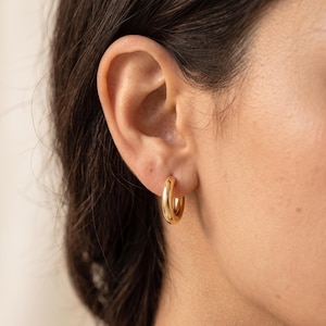 May include: A small, polished gold hoop earring with a thick, rounded profile. The earring features a smooth, reflective surface and a circular shape, securely fastened through the earlobe, showcasing a simple and classic metallic design.