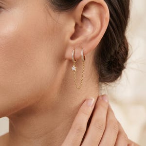 May include: Gold-coloured hoop earrings, each with small clear stones. One earring features a dangling star charm, connected to the other by a fine chain. The earrings are displayed on an ear.